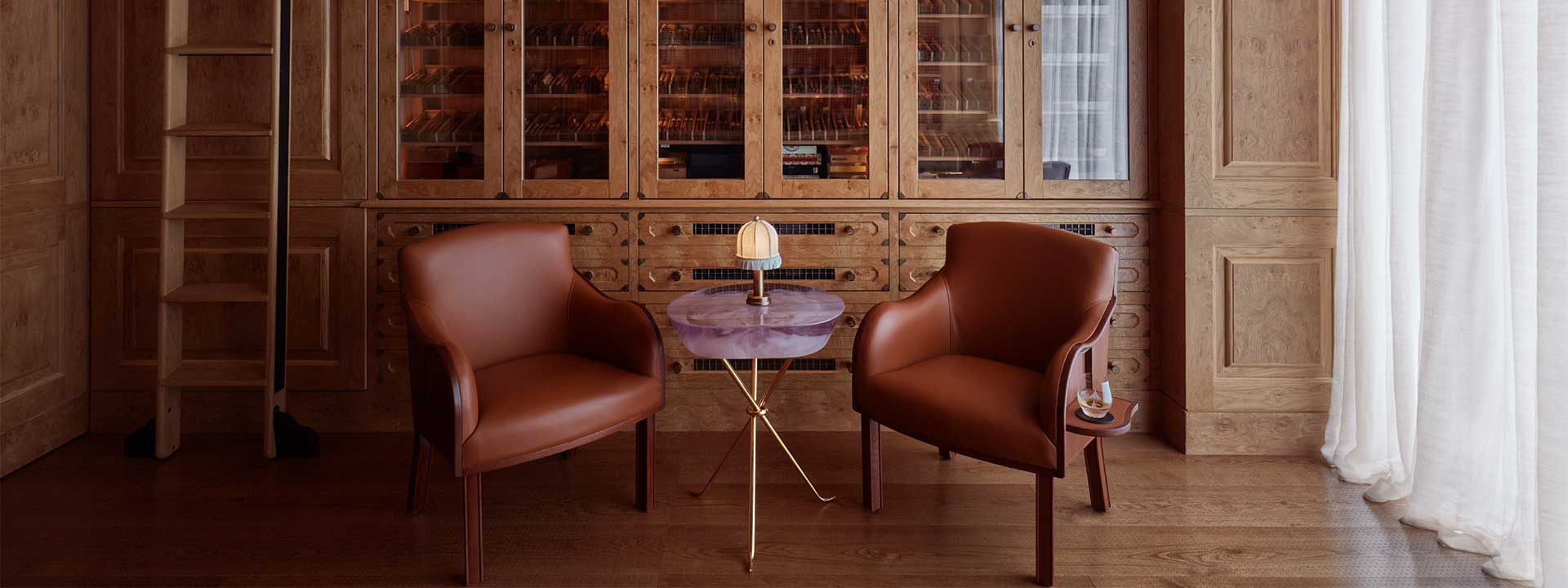 The Connaught Cigar Merchants inside view with a table and a lamp on it and two armchairs on each side of the table. On the back is the Cigar collection in the glass cupboard